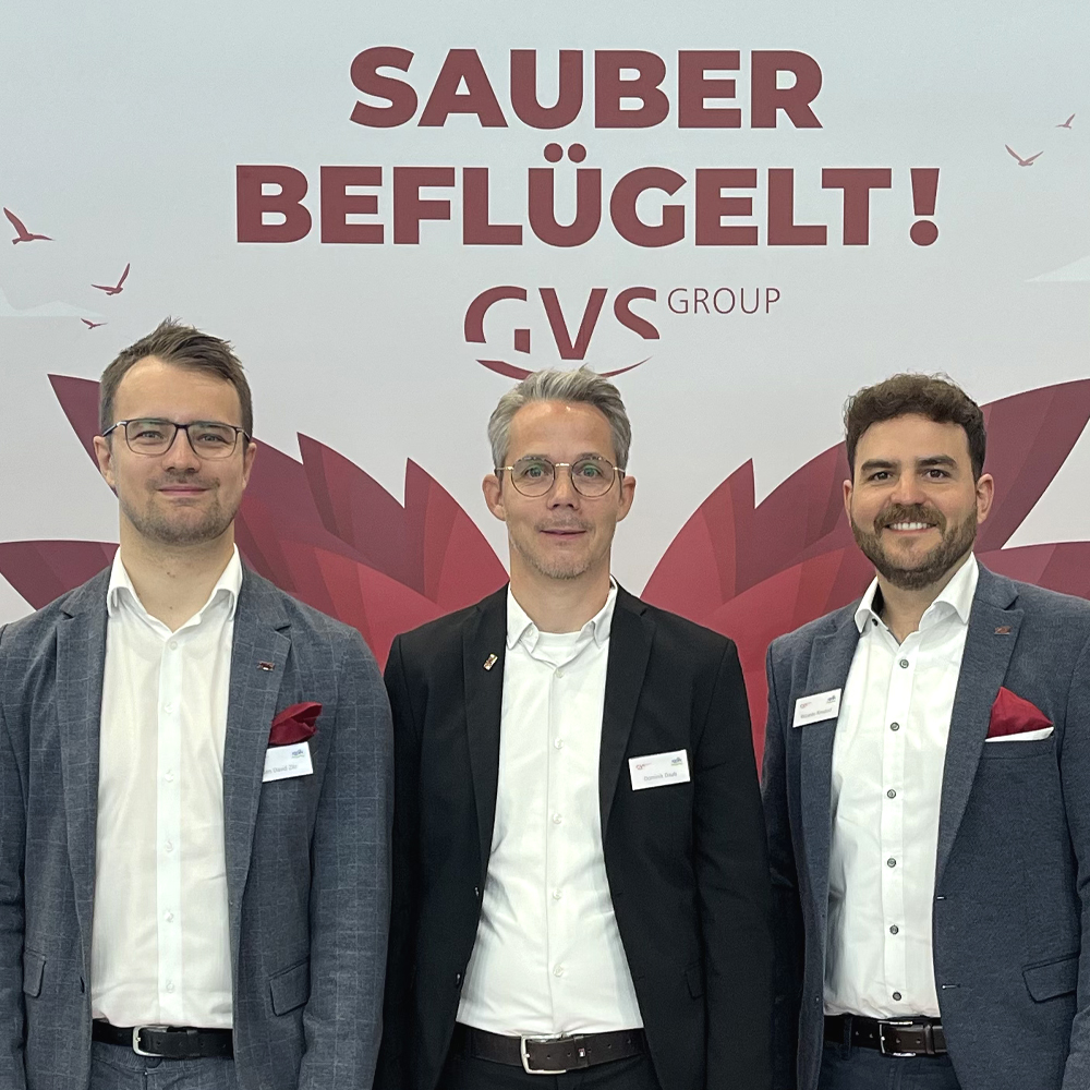 Drei Personen in Businesskleidung stehen vor einer Messewand mit dem Slogan „Sauber beflügelt!“ und dem Logo der GVS Group. Die Wand ist in Rot- und Weißtönen gestaltet, mit grafischen Elementen wie stilisierten Flügeln und Vögeln.
