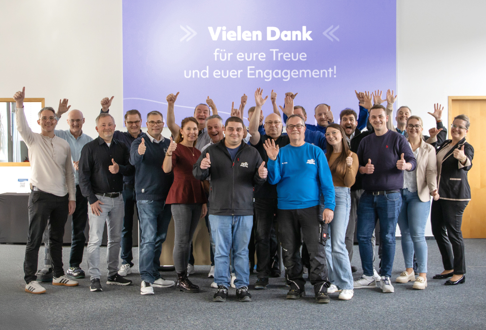 Gruppe von Mitarbeitenden steht vor einer großen Wand mit der Aufschrift „Vielen Dank für eure Treue und euer Engagement!“. Die Personen zeigen Daumen hoch und heben die Hände in die Luft.
