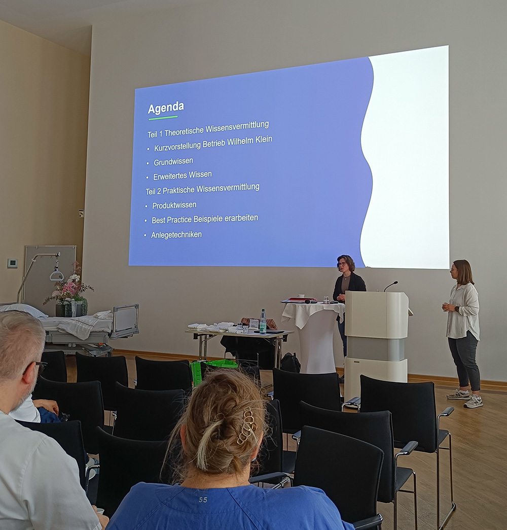 Schulungsveranstaltung in einem Seminarraum mit Präsentation auf großer Leinwand. Die Agenda zeigt Themen wie theoretische Wissensvermittlung, Produktwissen und Anlegetechniken.