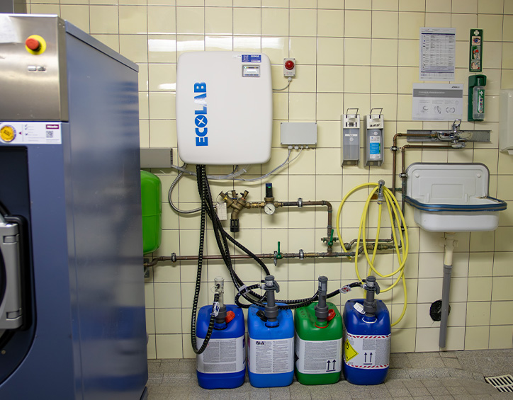 Dosiersystem für Wasch- und Reinigungschemie in einem professionellen Waschraum mit Chemikalienkanistern, Wandmodulen und Waschbecken.