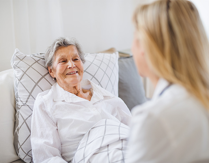 Pflegekraft spricht mit einer älteren Person, die im Bett liegt und von einer Decke mit grauem Karomuster bedeckt ist.